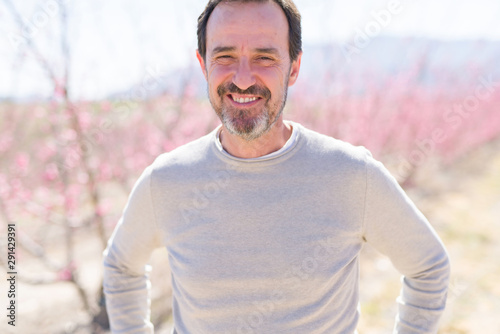 Handsome middle age senior man smiling cheerful on a peach trees garden with pink petals on a sunny day of spring