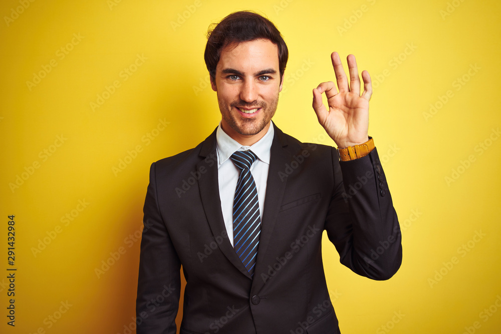Young handsome businessman wearing suit and tie standing over isolated yellow background smiling positive doing ok sign with hand and fingers. Successful expression. Young handsome businessman wearing suit and tie standing over isolated yellow background smiling positive doing ok sign with hand and fingers. Successful expression.