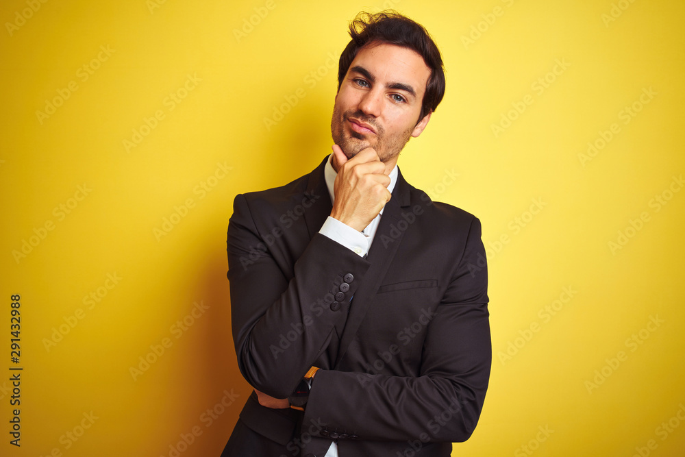 Young handsome businessman wearing suit and tie standing over isolated yellow background looking confident at the camera smiling with crossed arms and hand raised on chin. Thinking positive. Young handsome businessman wearing suit and tie standing over isolated yellow background looking confident at the camera smiling with crossed arms and hand raised on chin. Thinking positive.