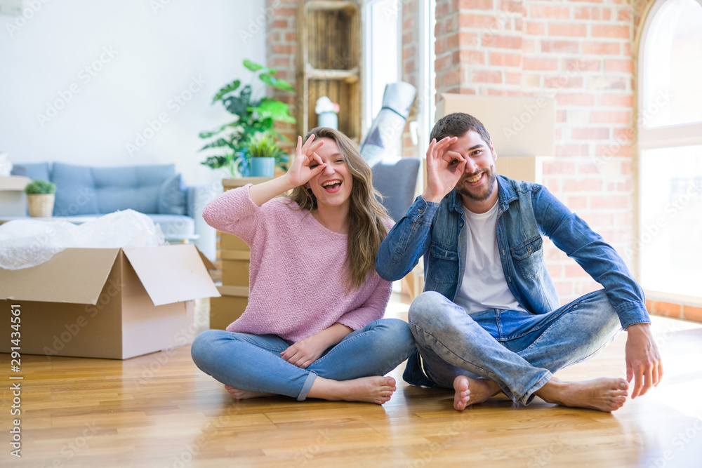 Young beautiful couple moving to a new house sitting on the floor doing ok gesture with hand smiling, eye looking through fingers with happy face.