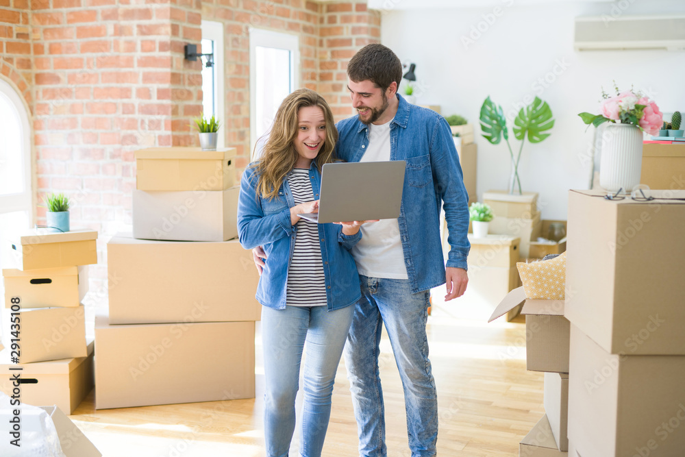 Young couple using computer laptop standing on a room around cardboard boxes, happy for moving to a new apartment Young couple using computer laptop standing on a room around cardboard boxes, happy for moving to a new apartment