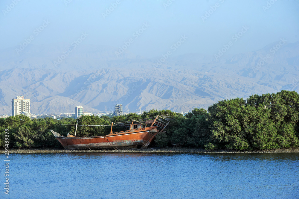 Panorama of Ras Al Khaimah, UAE .Lonely dhow traditional boat with sun lit mountains in the background. View from the corniche Panorama of Ras Al Khaimah, UAE .Lonely dhow traditional boat with sun lit mountains in the background. View from the corniche