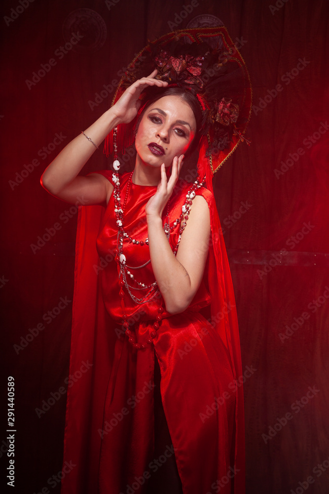 strict stylish girl in a fashionable long red dress and a voluminous hat on the background of smoke in the Studio at night alone. Halloween concept.