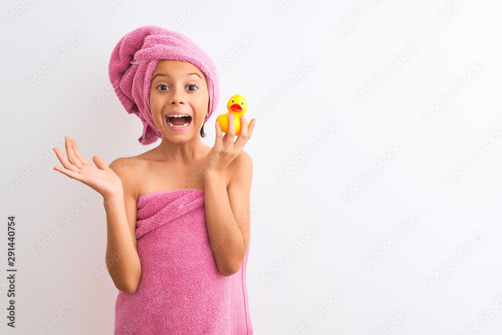 Beautiful child girl wearing shower towel holding duck over isolated white background very happy and excited, winner expression celebrating victory screaming with big smile and raised hands Beautiful child girl wearing shower towel holding duck over isolated white background very happy and excited, winner expression celebrating victory screaming with big smile and raised hands