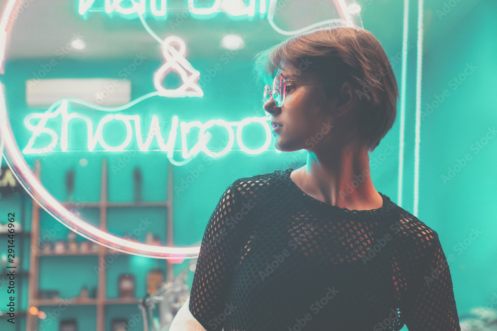 Hipster teen tomboy girl pretty face short hairstyle wear stylish glasses  on neon sign background, blue pink street light fashion trendy teenager  young woman profile view in glowing night city style Stock