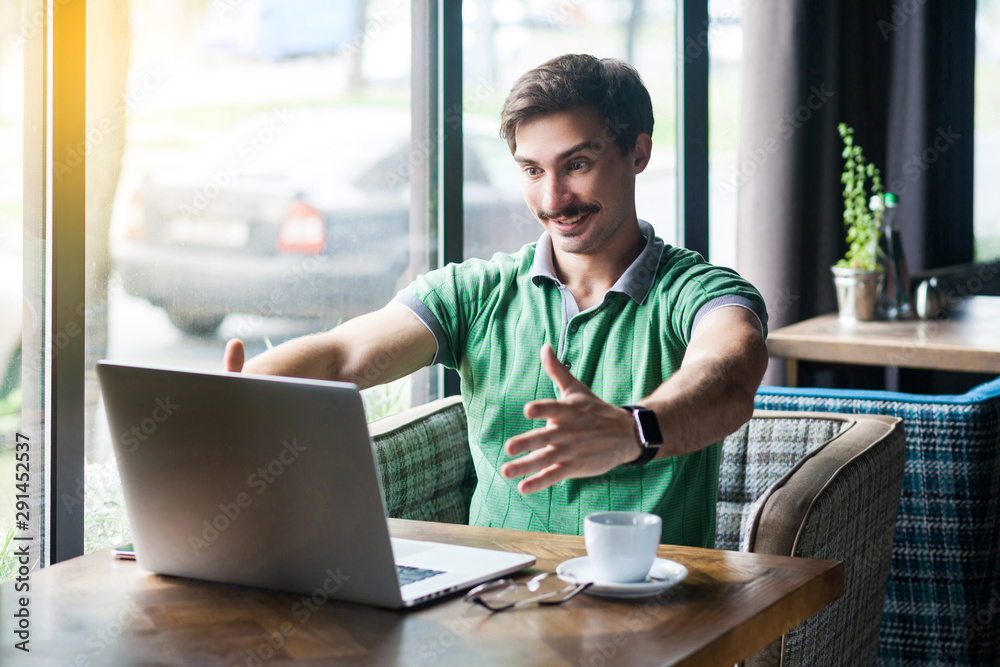 Give me hug. Young happy businessman in green t-shirt sitting and looking at laptop on video call and want to embrace and toothy smile. business concept. indoor shot near big window at daytime. Give me hug. Young happy businessman in green t-shirt sitting and looking at laptop on video call and want to embrace and toothy smile. business concept. indoor shot near big window at daytime.