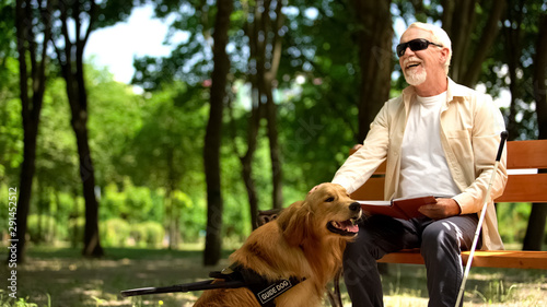 Cheerful blind man holding book and stroking assistance dog, enjoying life