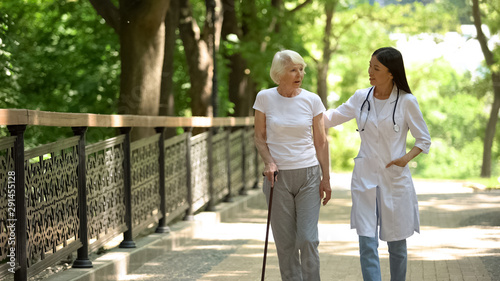 Doctor walking in park with elderly female patient with cane, rehabilitation