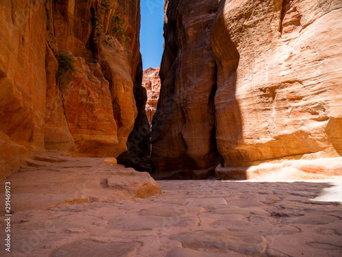 Steep cliffs of a canyon leading to the historical site of Petra, Jordan, light shining from blue sky on dark sandstone walls