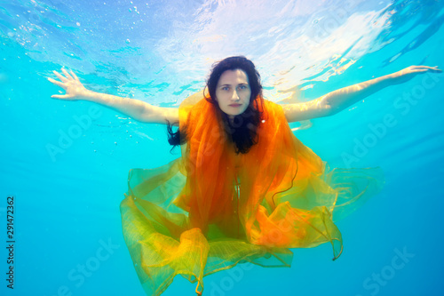 A beautiful girl underwater with a yellow cloth looks and poses for the camera on a Sunny day, arms outstretched, against the sky. Fashion portrait.