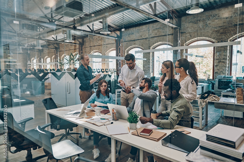 Group of young modern people in smart casual wear communicating and using modern technologies while working in the office