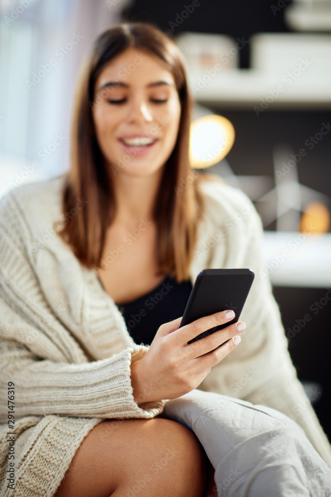 Beautiful smiling caucasian brunette sitting dressed in beige sweater sitting on bed in bedroom and using smart phone. Selective focus on hand. Beautiful smiling caucasian brunette sitting dressed in beige sweater sitting on bed in bedroom and using smart phone. Selective focus on hand.