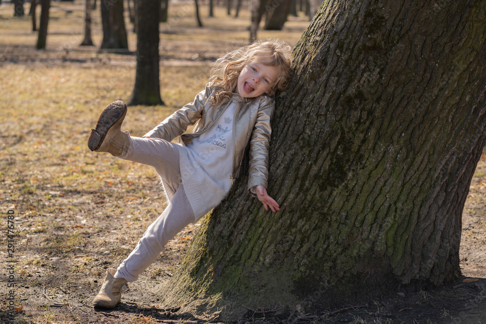 pretty curly kid girl in leather jacket posing near big tree in the city park alone pretty curly kid girl in leather jacket posing near big tree in the city park alone