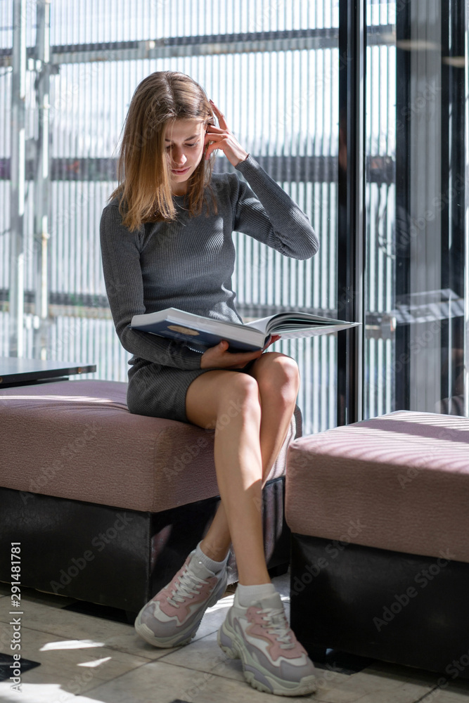 European student girl reading book while sitting on sofa near large window in public library European student girl reading book while sitting on sofa near large window in public library