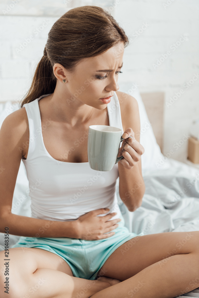stressed young woman sitting on bed, touching belly and holding cup of tea