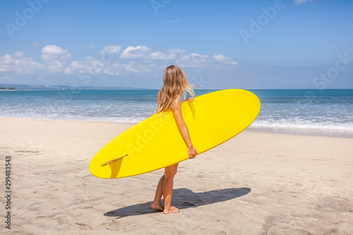 Back view of young woman with long hair holding the surfboard preparing to surf on the beach with sea on background on Bali island, Indonesia. Active lifestyle. Holiday leisure. Blue sky.
