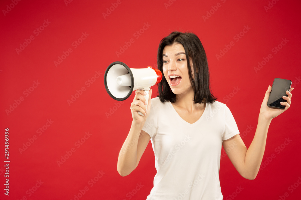 Caucasian young woman's half-length portrait on red studio background. Beautiful female model in shirt. Concept of human emotions, facial expression, sales. Holding phone and calling in mouthpeace. Caucasian young woman's half-length portrait on red studio background. Beautiful female model in shirt. Concept of human emotions, facial expression, sales. Holding phone and calling in mouthpeace.