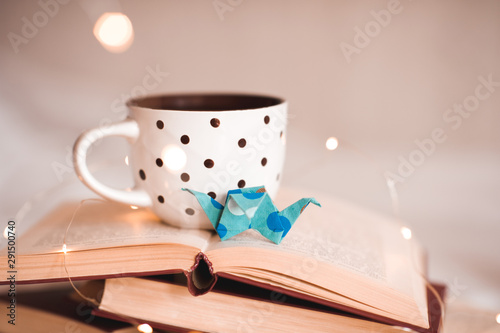 Cup of tea with paper origami crane on open books close up over lights. Good morning.