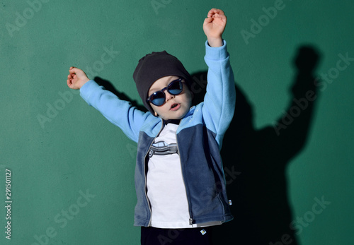 Portrait of cool kid boy in blue sunglasses, hat, fleece jacket and white t-shirt having fun jumping dancing on green