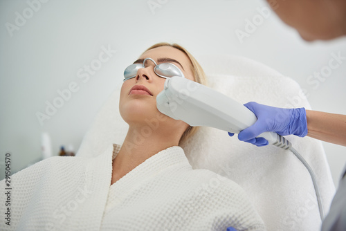 Young lady in goggles receiving laser facial treatment in beauty salon