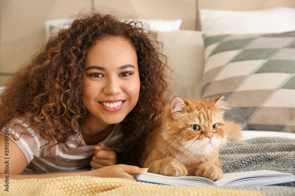 Young African-American woman with cute cat reading book at home - obrazy, fototapety, plakaty 