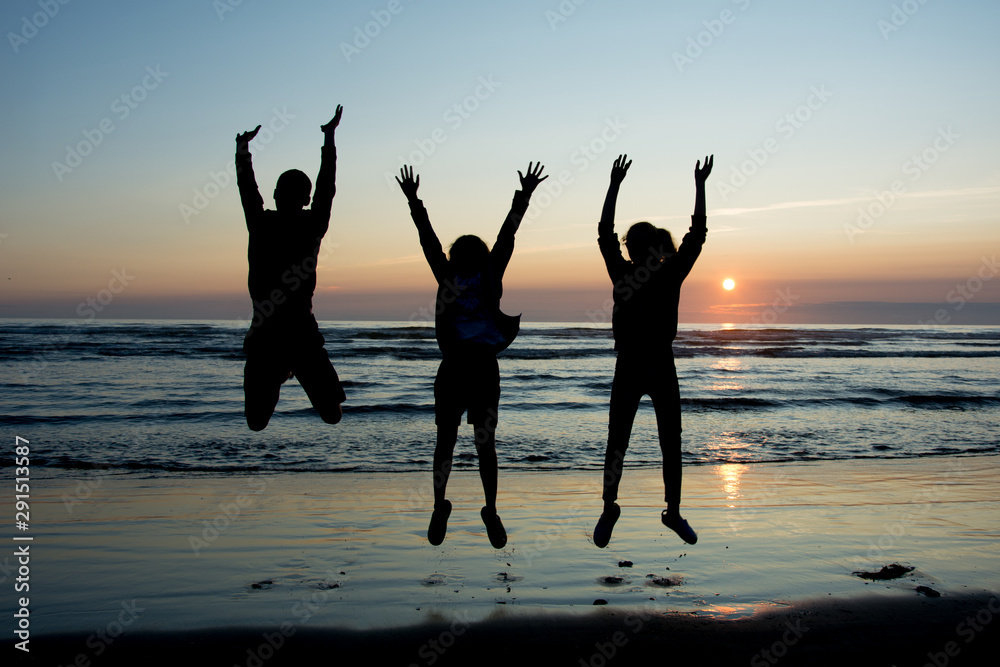 Fototapeta premium Silhouettes of three teanagers jumping out joyfully on the beach with setting sun at the bakcground