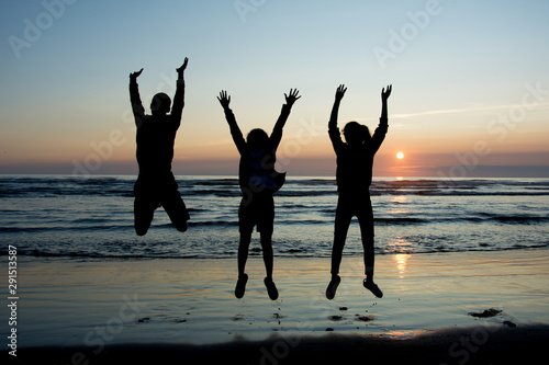 Silhouettes of three teanagers jumping out joyfully on the beach with setting sun at the bakcground