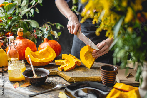 Woman cook cuts an orange pumpkin with a knife into slices for baking. Concept autumn food in a cozy dark kitchen