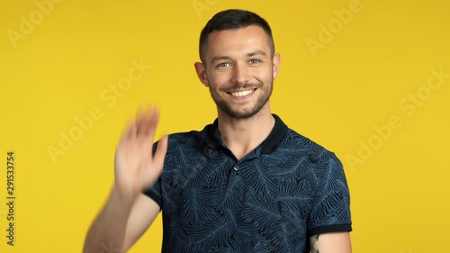 Handsome happy man waving hello or goodbye to camera on yellow background photo