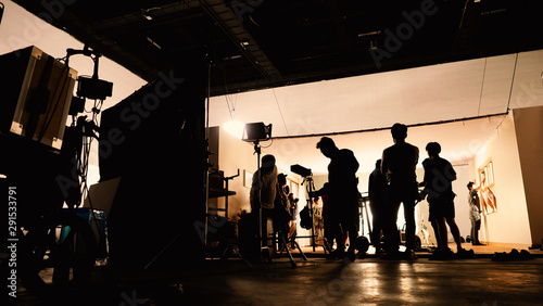 Behind the shooting video production and lighting set for filming which movie crew team working and silhouette shadow of camera and professional equipment in big studio for commercial advertising.