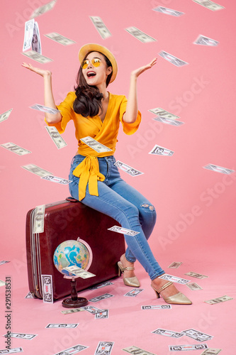 Smiling woman traveler sitting on luggage raising arms and money in holiday on pink backgrounds, relaxation concept, travel concept