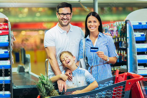 Happy family with child and shopping cart buying food at grocery store or supermarket, woman holding credit card.