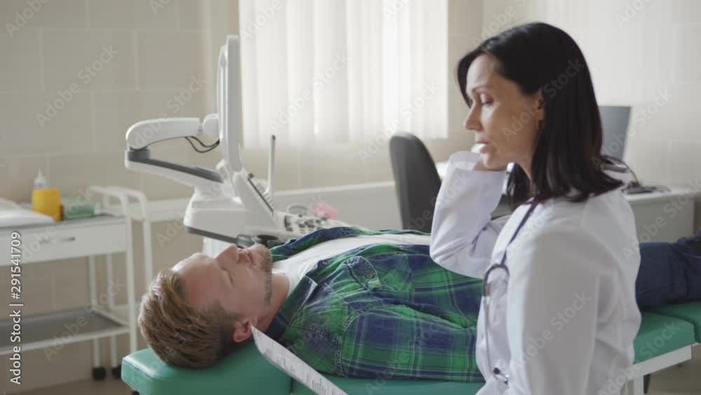 Tracking medium shot of middle aged male patient lying on couch in doctors office and talking to female physician reading his medical history Tracking medium shot of middle aged male patient lying on couch in doctors office and talking to female physician reading his medical history