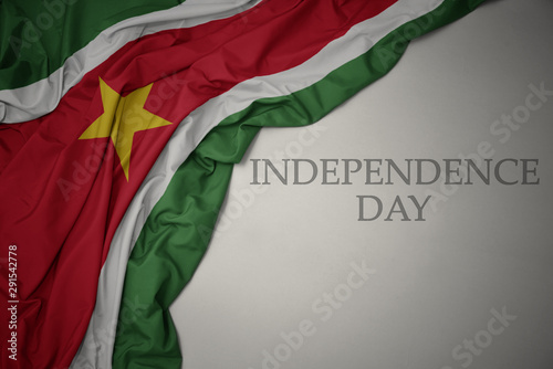 waving colorful national flag of suriname on a gray background with text independence day.