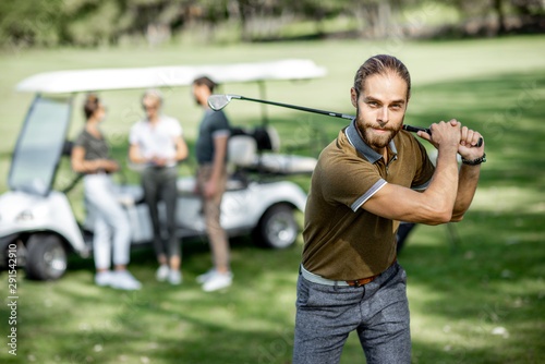 Portrait of a handsome man swinging with putter, playing golf on the course with friends and golf car on the background on a sunny weather