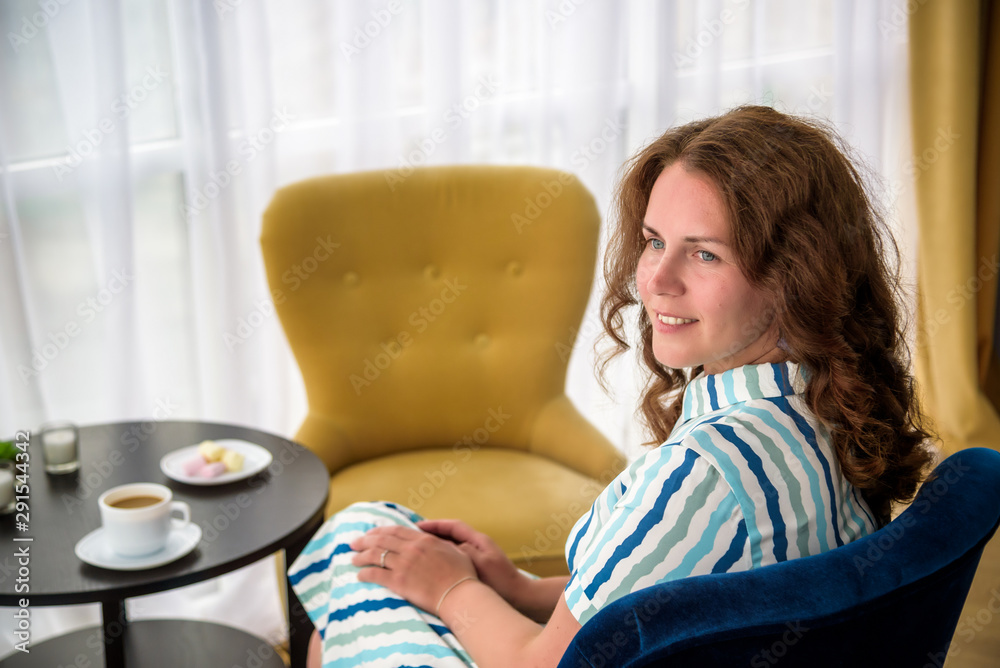 Young woman at home sitting on modern chair in front of window relaxing in her living room and drinking coffee or tea with sweets or desert. Relax before new business day Young woman at home sitting on modern chair in front of window relaxing in her living room and drinking coffee or tea with sweets or desert. Relax before new business day