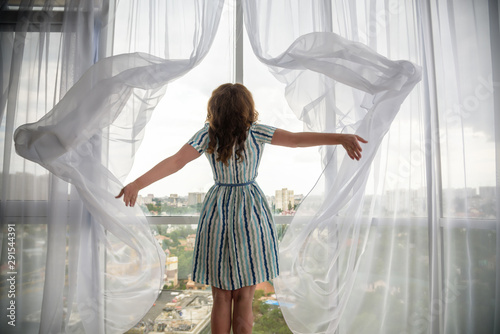 Konstfotografi Rear view of a young joyful woman wearing fashion dress and holding the curtains