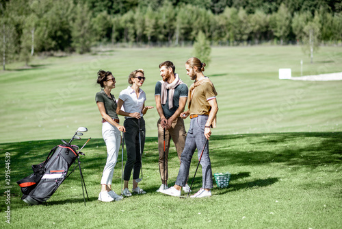 Young and elegant friends standing together with golf equipment, having fun during a golf play on the beautiful course on a sunny day