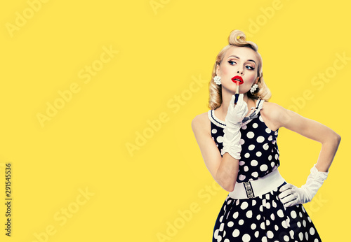 Young woman in pin up style black and white dress in polka dot, applying lipstick, isolated over yellow color background