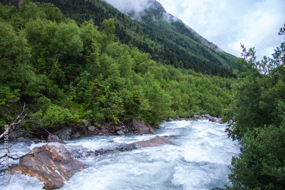 Beautiful panorama of the Caucasus mountains. Top of mountain range is covered in fog. Clouds lie on mountains. Fast mountain river. Background image for travel and nature. Beautiful panorama of the Caucasus mountains. Top of mountain range is covered in fog. Clouds lie on mountains. Fast mountain river. Background image for travel and nature.