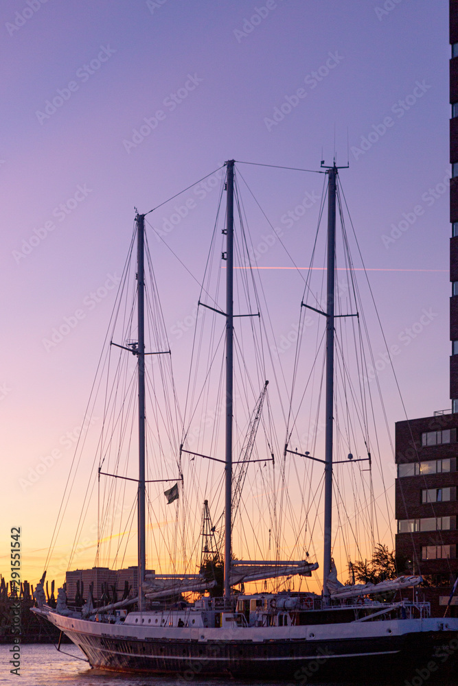 Docked traditional sailboat next to high rise building against a deep purple and orange sunset sky