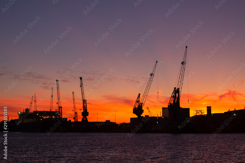 Silhouette of a shipyard with cranes at golden hour orange, purple and red colours in the background