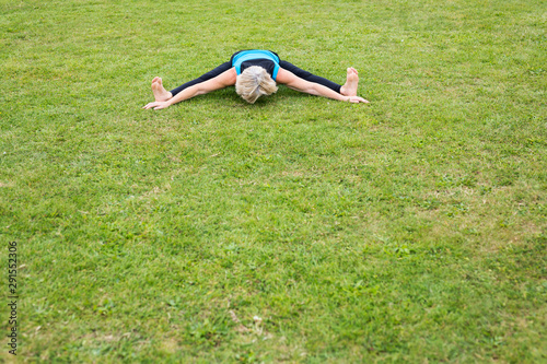 A middle aged woman practicing yoga barefoot outside in a grassy park. She is wearing a bright blue vest and black leggings. The style of yoga she is doing is Hatha Yoga