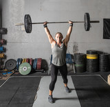 Strong young woman lifting heavy barbell over her head in gym.