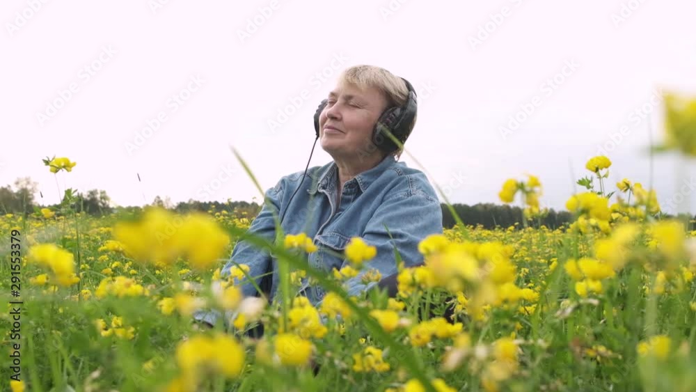 Happy relaxed mature woman sits with her eyes closed wearing headphones and listen music on field, around her wind-moved flowers and insects fly. Sonic Therapy. Sensitivity to nature. Slow motion. Happy relaxed mature woman sits with her eyes closed wearing headphones and listen music on field, around her wind-moved flowers and insects fly. Sonic Therapy. Sensitivity to nature. Slow motion.