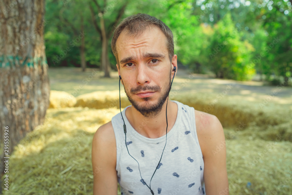 A young handsome man with beard in a t-shirt is sits in the hay, listens to music and sings in the park. A young handsome man with beard in a t-shirt is sits in the hay, listens to music and sings in the park.