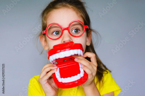 Little funny girl kid with toothbrush, dental mockup (jaw), red glasses in hand. Concept of health, oral hygiene, people and beauty. Space for your text. Selective focus.