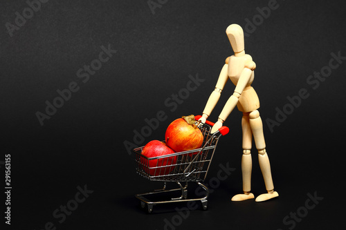 Conceptual image of a miniature shopping trolley filled with apples being pushed by a manikin isolated on a black background with space for copy photo