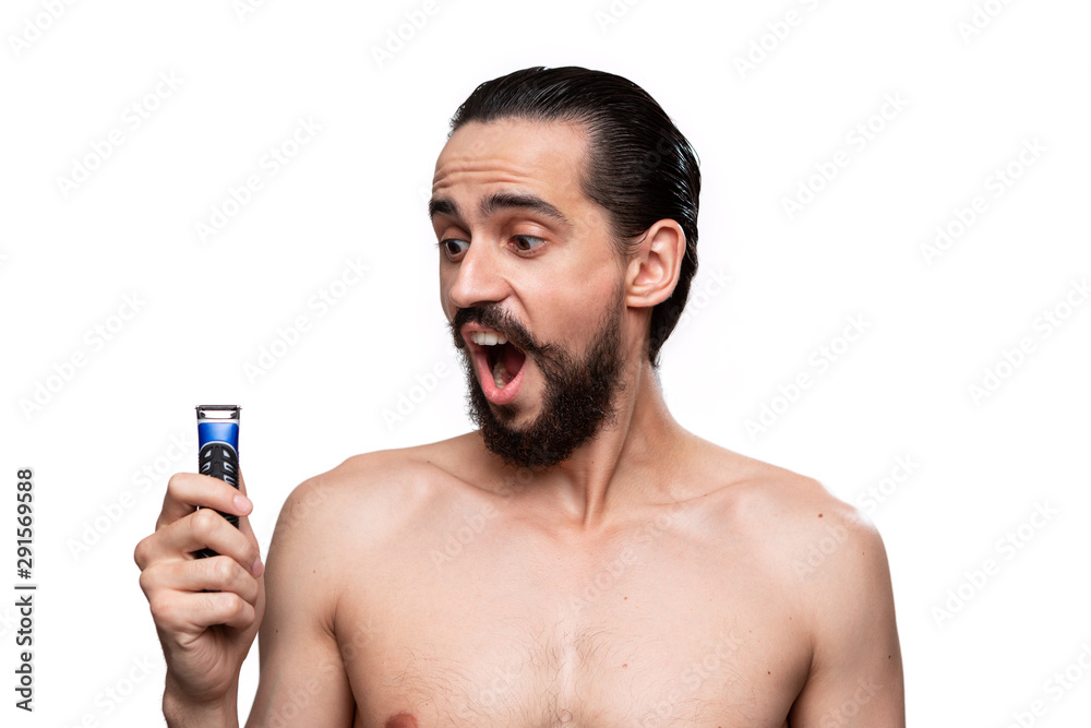 Fototapeta premium Handsome bearded man with mustache holds electric shaver standning bare isolated over white background. Concept of morning treatment and shaving. Time to trim your beard. Morning routine