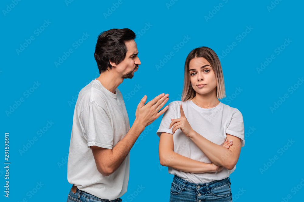 Brunet bearded man with mustaches in white t-shirt and blue jeans asking his girlfriend for forgiveness isolated over blue background. Concept of guilt. Brunet bearded man with mustaches in white t-shirt and blue jeans asking his girlfriend for forgiveness isolated over blue background. Concept of guilt.